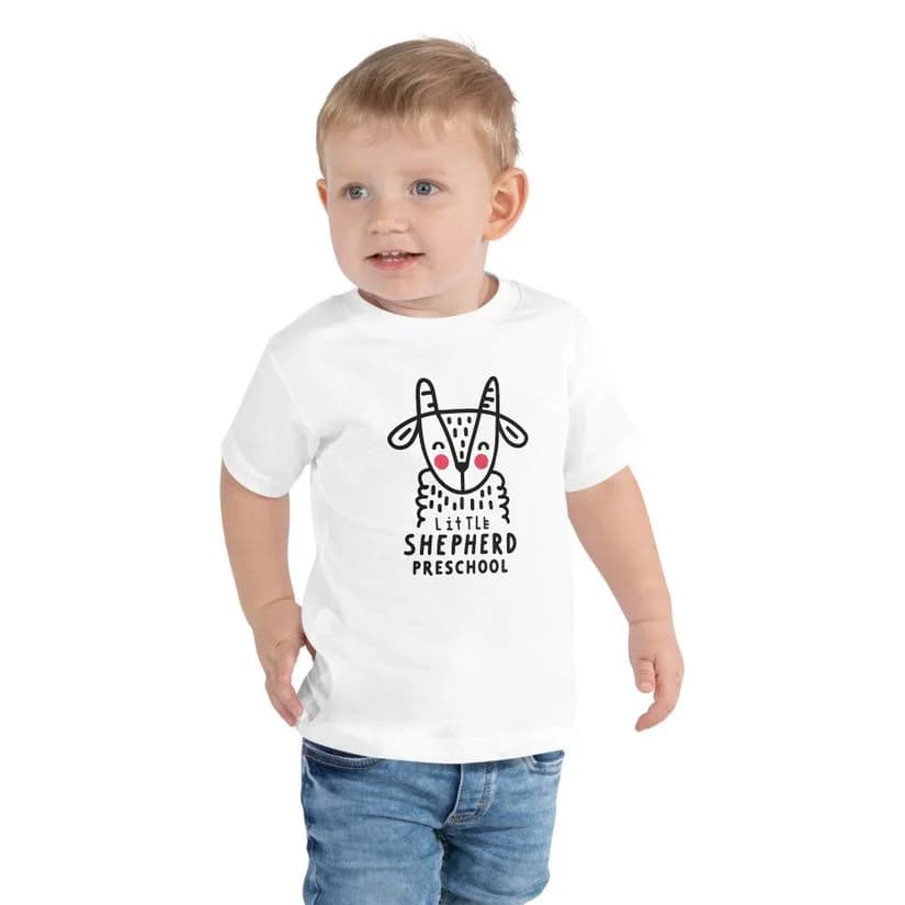 A toddler wearing a white Little Shepherd Preschool t-shirt with a sheep graphic.